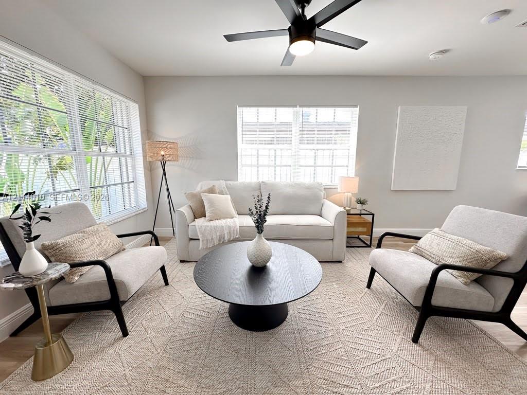 Living area featuring light-toned walls, wood-finish flooring, dual-pane windows with horizontal blinds, and a central ceiling fan with integrated lighting