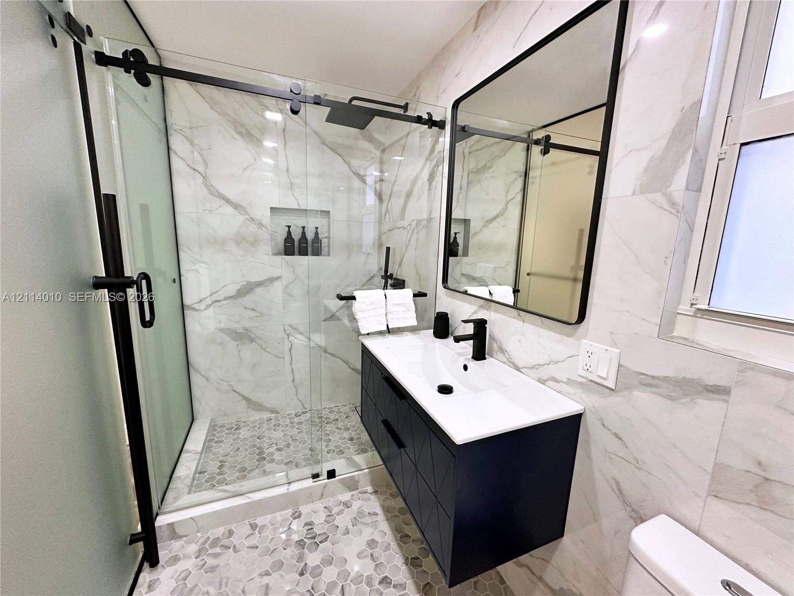Walk-in shower featuring a sliding glass door, matte black hardware, integrated wall niche, and stone-tiled floor with a matching vanity and black-framed mirror