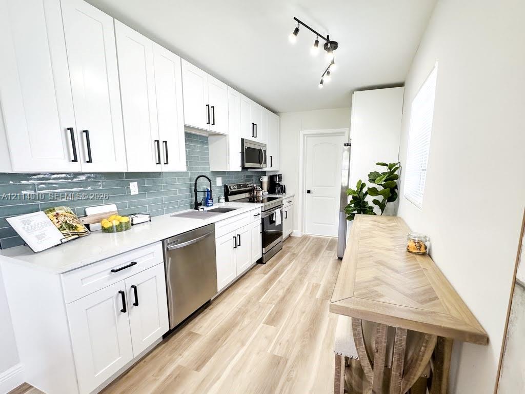 Kitchen featuring white shaker-style cabinetry, light-toned countertop, blue glass tile backsplash, stainless steel appliances, and wood-finish flooring
