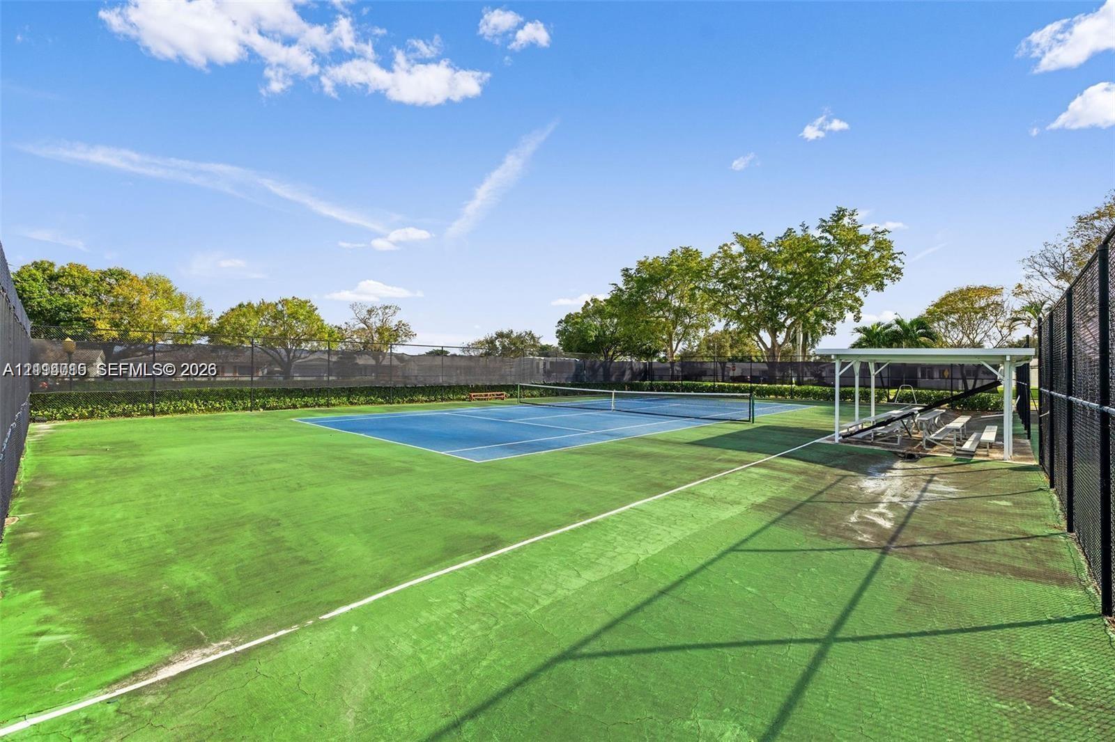 Tennis court with a blue hard-surface center, perimeter chain-link fencing, and a covered spectator area