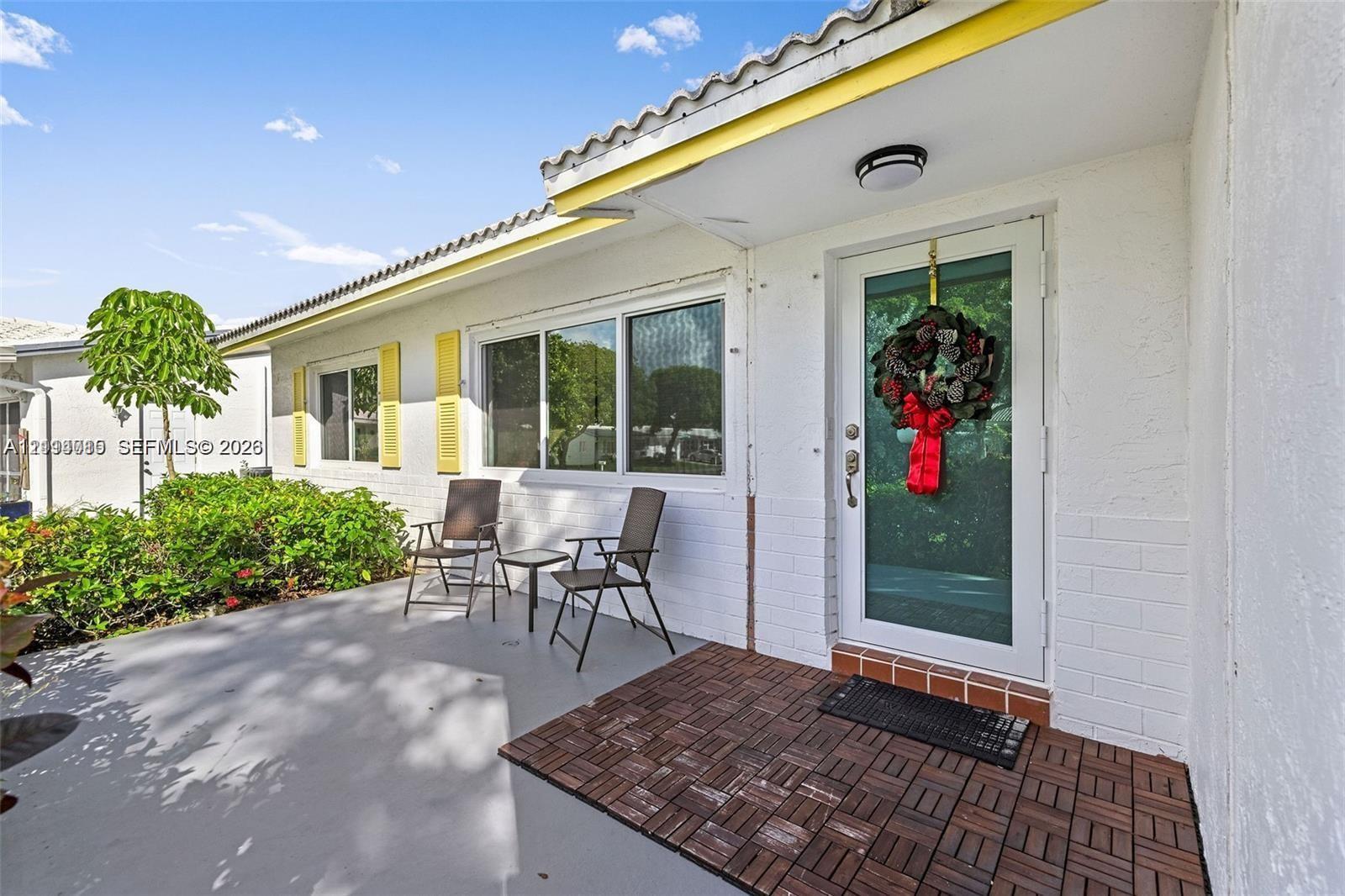 Exterior entryway featuring white painted brick masonry, yellow louvered shutters, decorative tile flooring, and a glass-paneled entry door