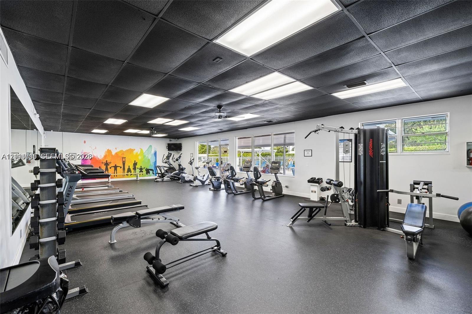 Fitness area featuring a dark rubberized floor, recessed panel ceiling, multiple rows of inset overhead lighting, and wall-to-wall mirrors