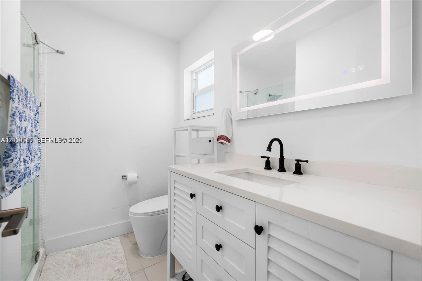 White vanity with louvered cabinet doors, stone-finish countertop, matte black widespread faucet, integrated LED mirror, and glass-enclosed shower