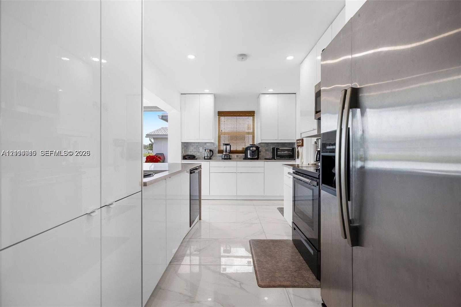 Kitchen featuring glossy white flat-panel cabinetry, light-toned stone-finish tile flooring, a stone-tile backsplash, stainless steel appliances, and recessed lighting