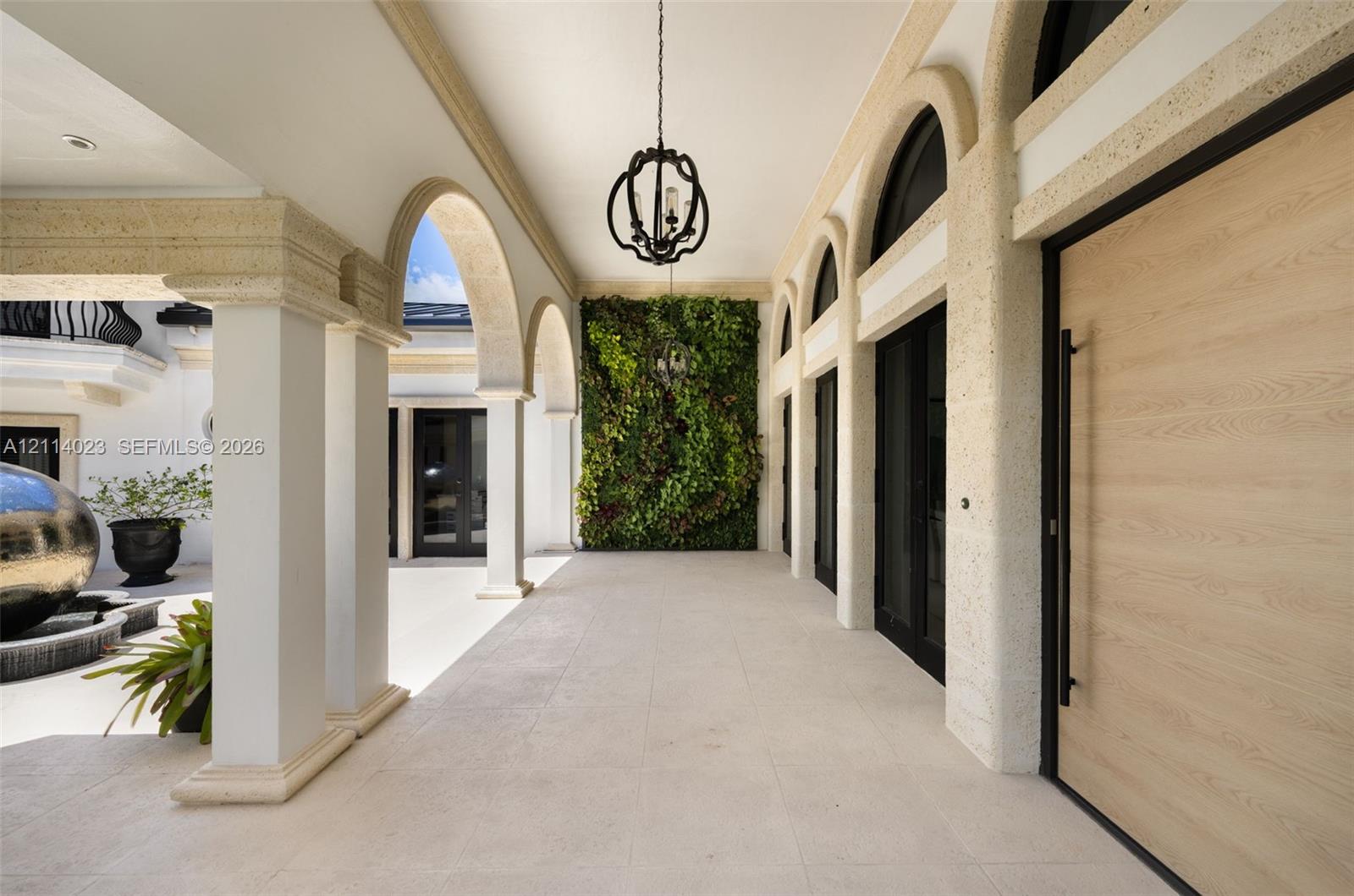 Covered loggia featuring arched architectural columns, stone-textured detailing, a large-format light-toned tile floor, a decorative hanging pendant fixture, and a vertical green garden wall