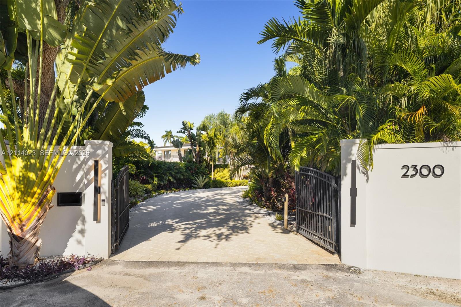 Gated entrance featuring decorative stucco walls, iron swing gates, paver driveway, and mature tropical landscaping Gated entrance featuring decorative stucco walls, iron swing gates, paver driveway, and mature tropical landscaping