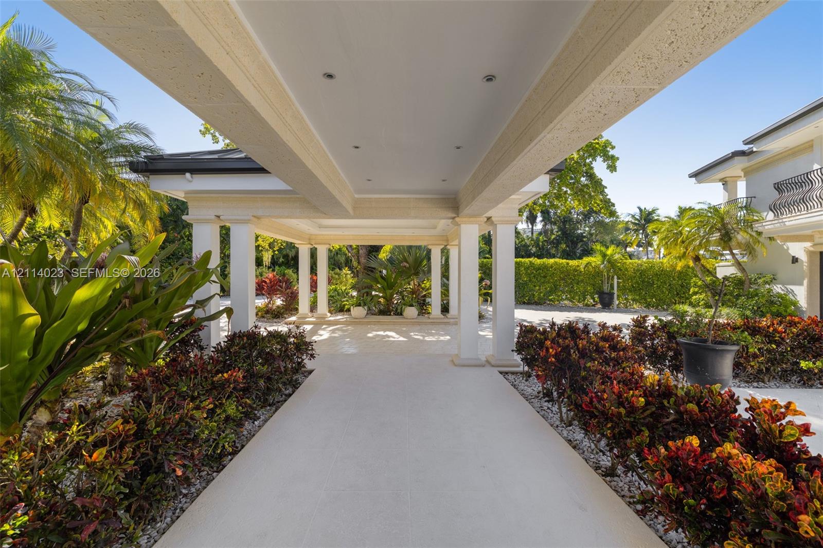 Covered porte-cochre featuring a light-toned stone tile surface, structural white columns, recessed lighting, and surrounding professional landscaping Covered porte-cochre featuring a light-toned stone tile surface, structural white columns, recessed lighting, and surrounding professional landscaping