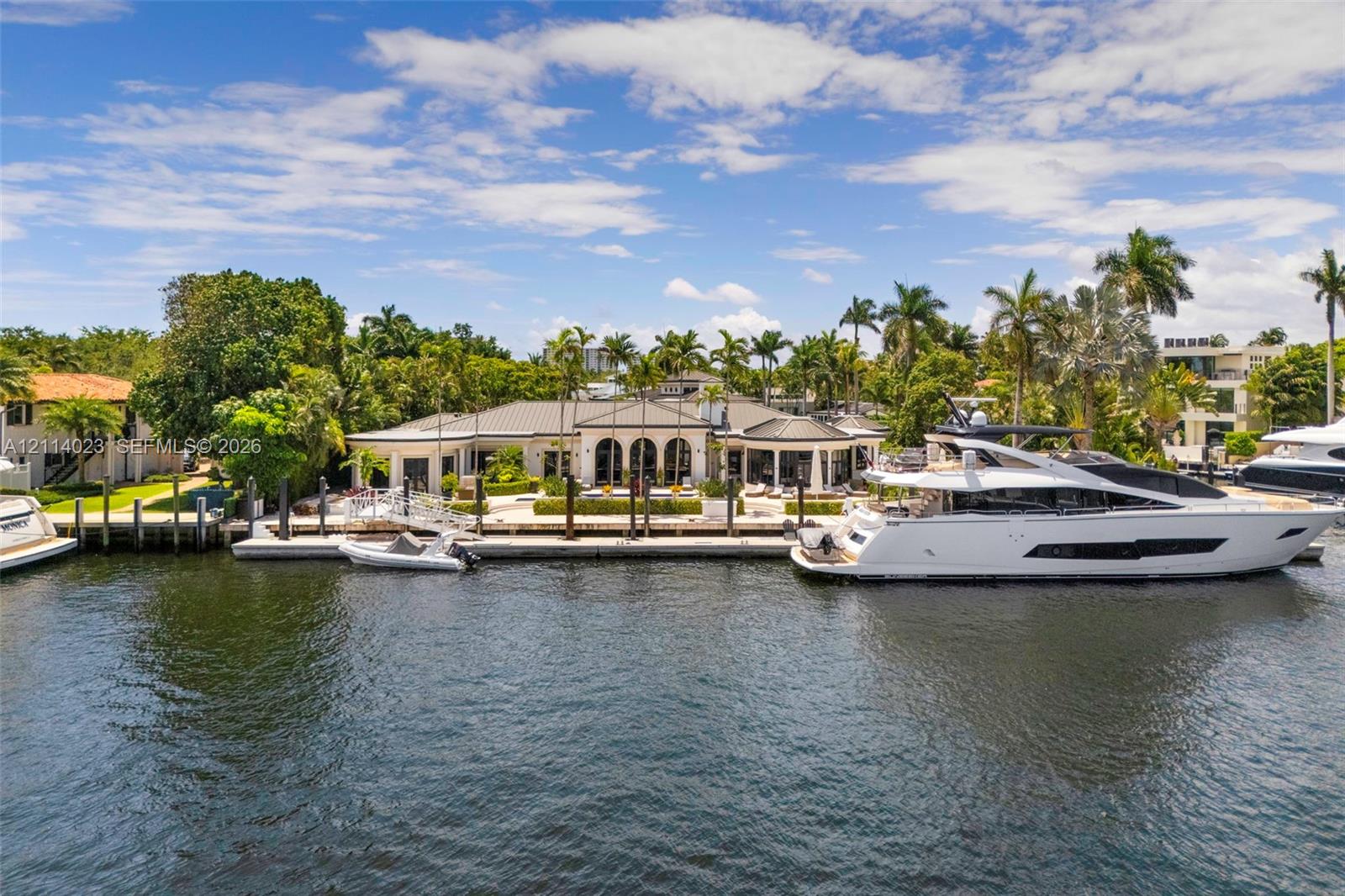 Waterfront estate featuring a deep-water dock, expansive stone patio, arched architectural details, and mature tropical landscaping