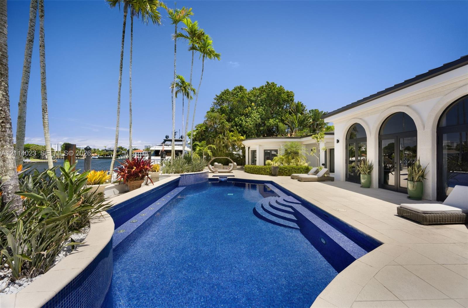 Waterfront patio featuring a tiled swimming pool, stone-finish decking, arched glass entryways, and manicured tropical landscaping