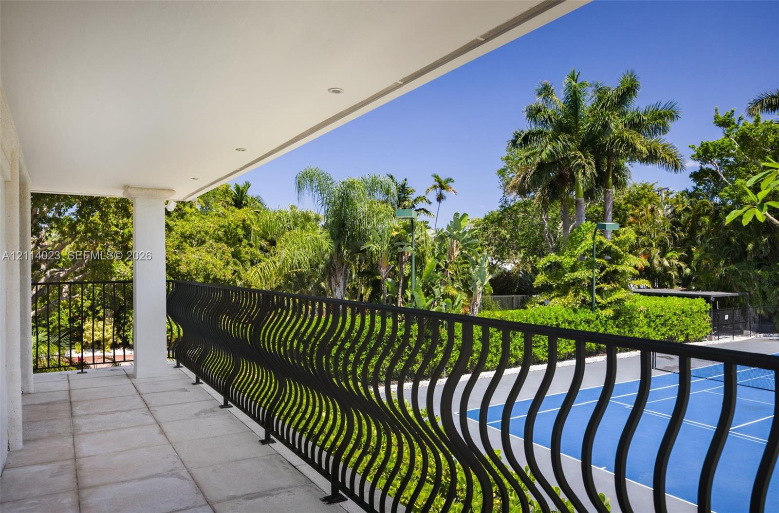 Covered balcony featuring stone-tile flooring, recessed lighting, and black metal wavy-pattern railing overlooking a hard-surface sport court and mature landscaping