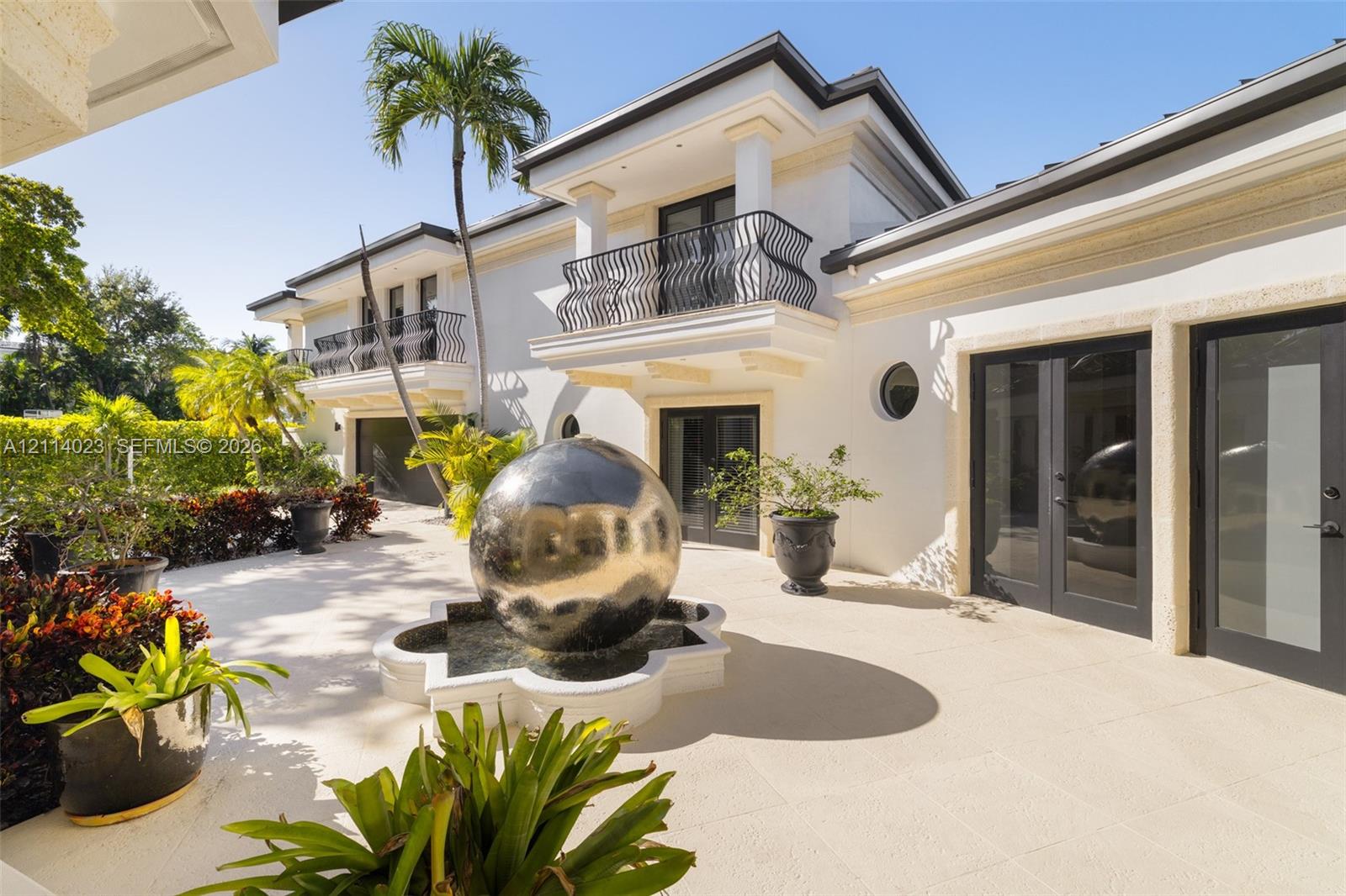 Exterior patio featuring stone-tiled flooring, a central sphere water feature, wrought-iron balcony railings, and dual-story structure with white stucco finish