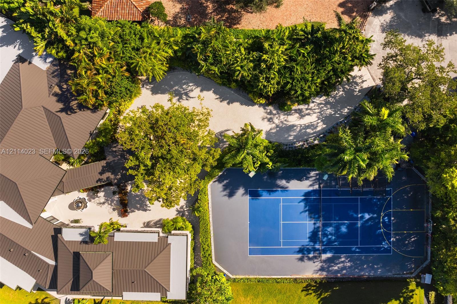 Aerial perspective of the estate featuring a multi-sport court with blue hard surfacing, expansive stone-paved driveway, metal roofing, and mature landscape canopy Aerial perspective of the estate featuring a multi-sport court with blue hard surfacing, expansive stone-paved driveway, metal roofing, and mature landscape canopy