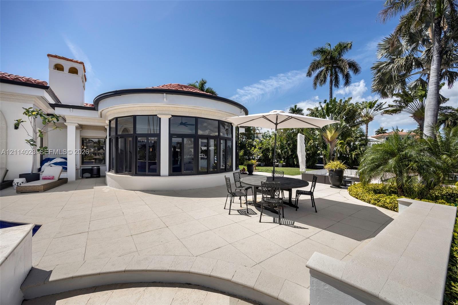 Expansive stone patio featuring a curved architectural structure, multi-pane floor-to-ceiling windows, integrated masonry seating, and a terracotta tile roof