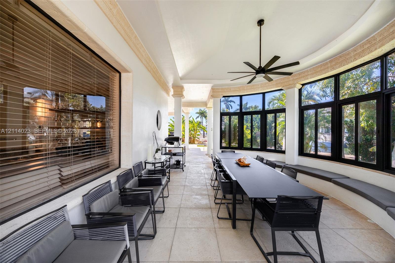 Covered outdoor patio featuring light-toned tile flooring, curved architecture, floor-to-ceiling windows with black frames, built-in bench seating, and a ceiling fan
