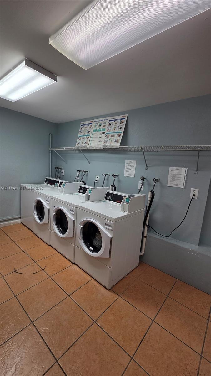 Laundry area featuring ceramic tile flooring, three white front-load washers, and a wall-mounted wire storage shelf Laundry area featuring ceramic tile flooring, three white front-load washers, and a wall-mounted wire storage shelf