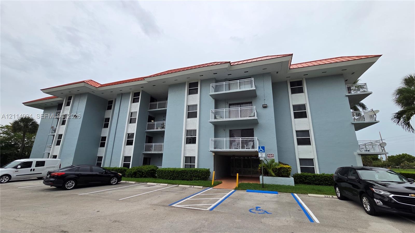 Multi-story residential building featuring a red metal roof, stucco exterior, stacked private balconies with railings, and paved surface parking