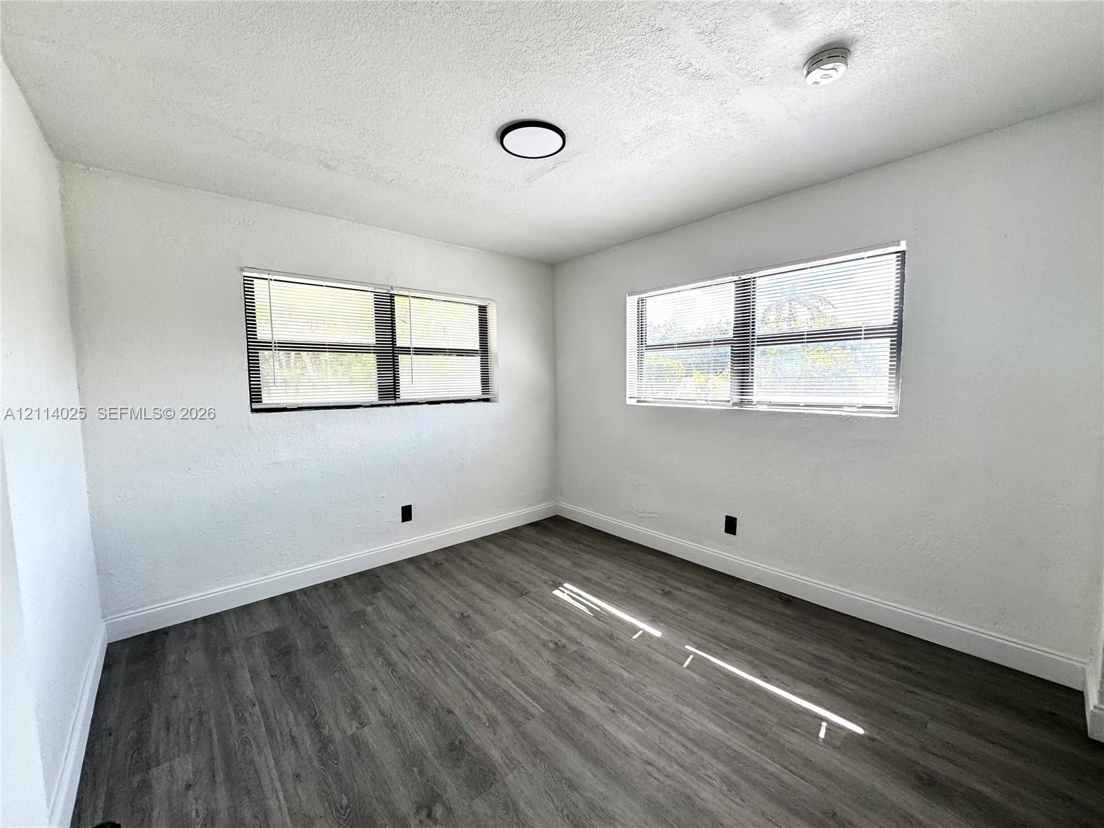 Room featuring wood-finish flooring, white walls, dual windows with integrated blinds, and flush-mount ceiling lighting