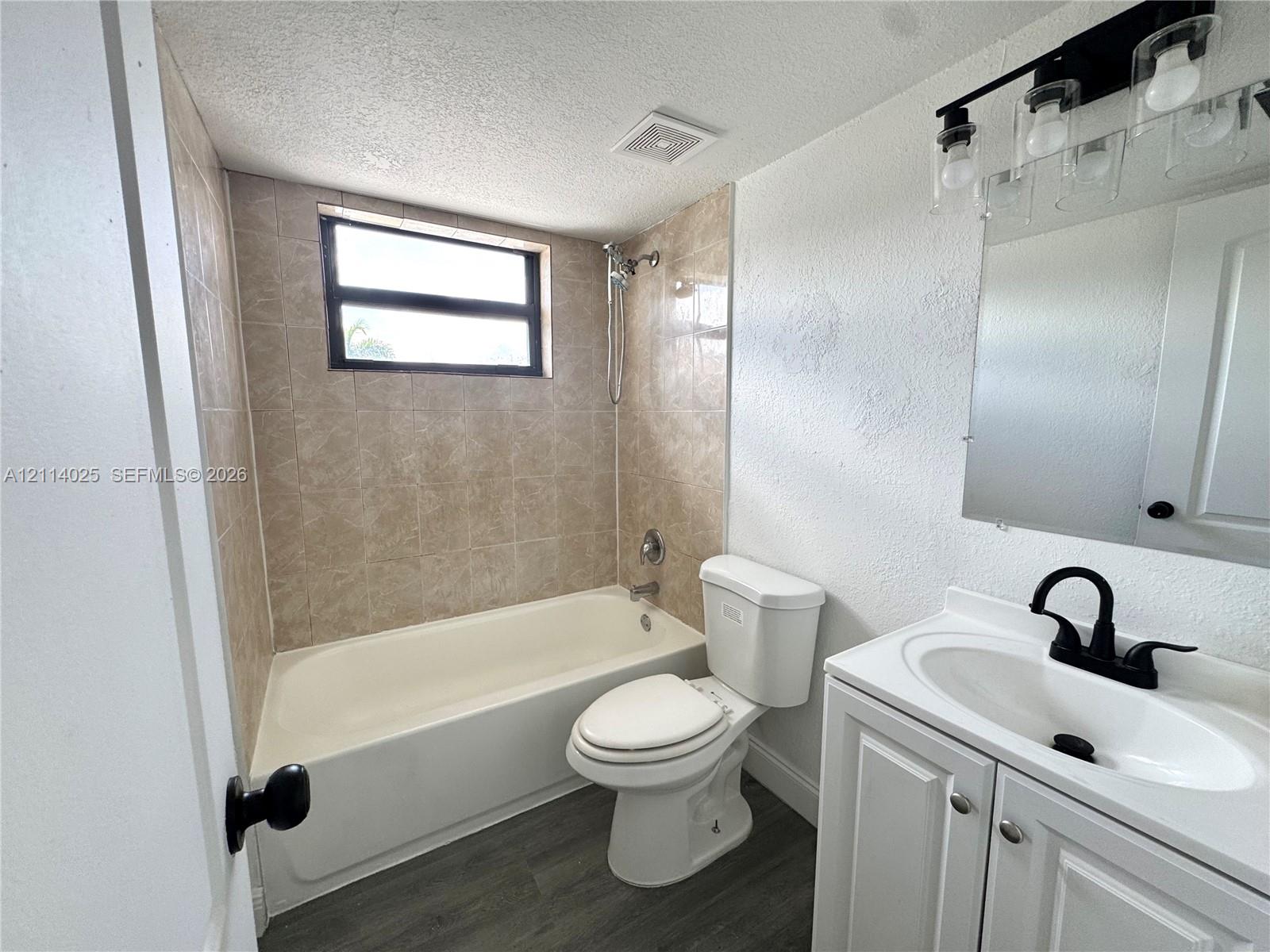 Bathroom featuring a white vanity cabinet with a integrated sink, matte black faucet, and contemporary light fixture, complemented by wood-finish flooring and a tiled shower surround