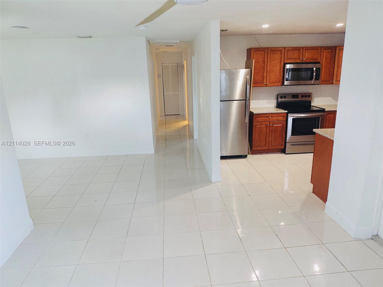 Kitchen featuring wood-finish cabinetry, neutral solid-surface countertops, and stainless steel appliances, set against white tile flooring Kitchen featuring wood-finish cabinetry, neutral solid-surface countertops, and stainless steel appliances, set against white tile flooring