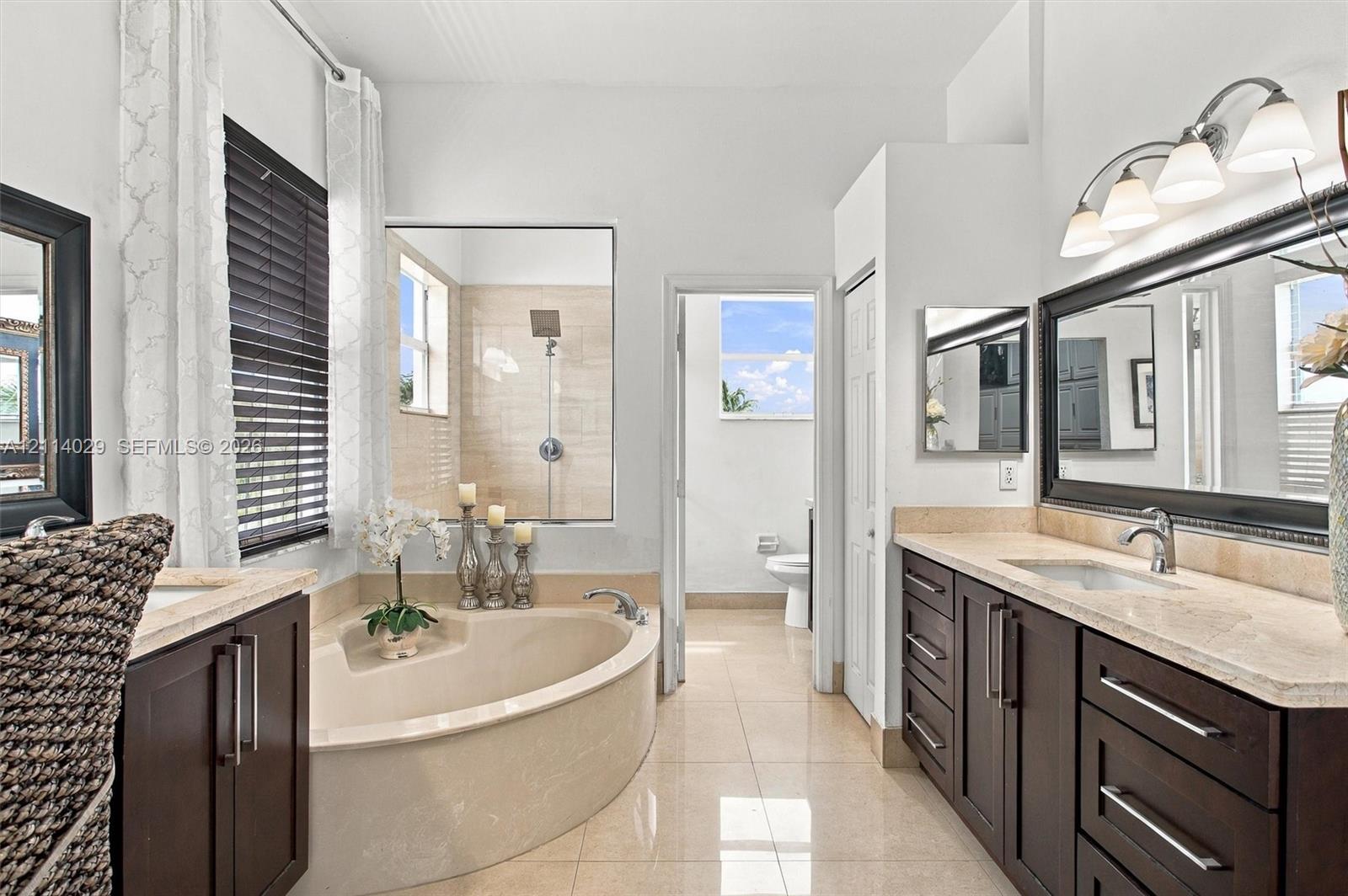 Dual-vanity cabinetry with stone-finish countertops, corner soaking tub, large-format tile flooring, walk-in shower with tiled surround, and multi-light vanity fixtures