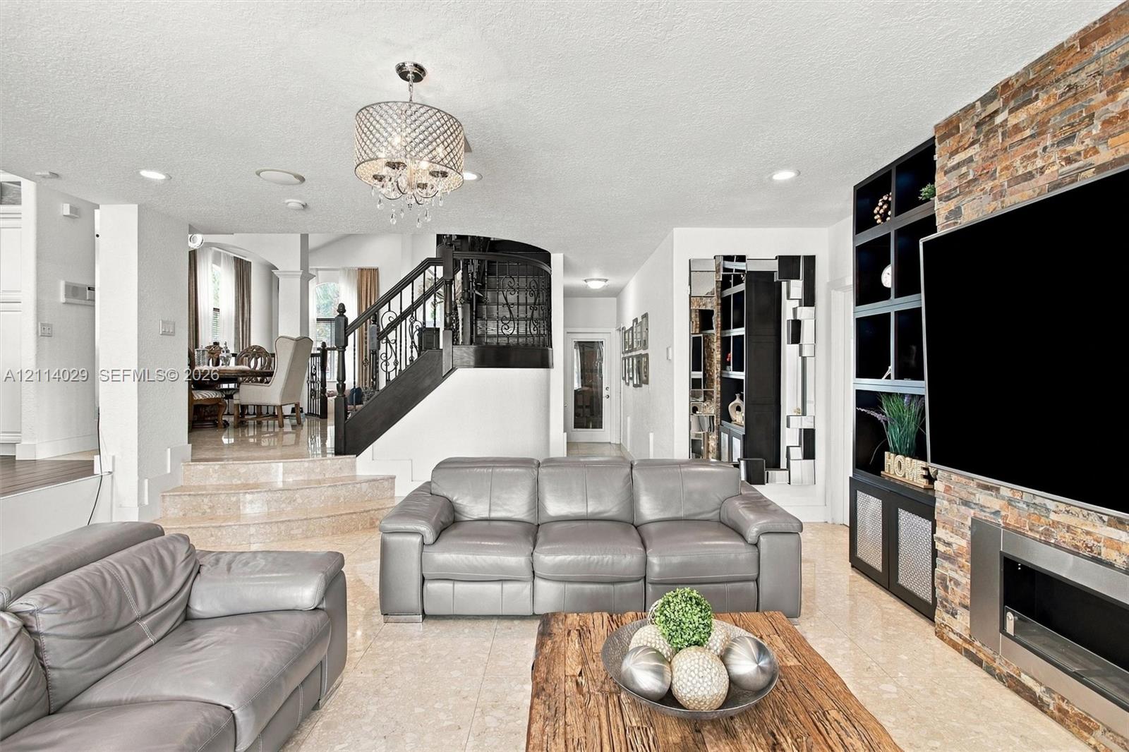 Living area featuring tile flooring, a floor-to-ceiling stone-clad fireplace with built-in dark cabinetry, a multi-tiered staircase with wrought iron railing, and a decorative chandelier