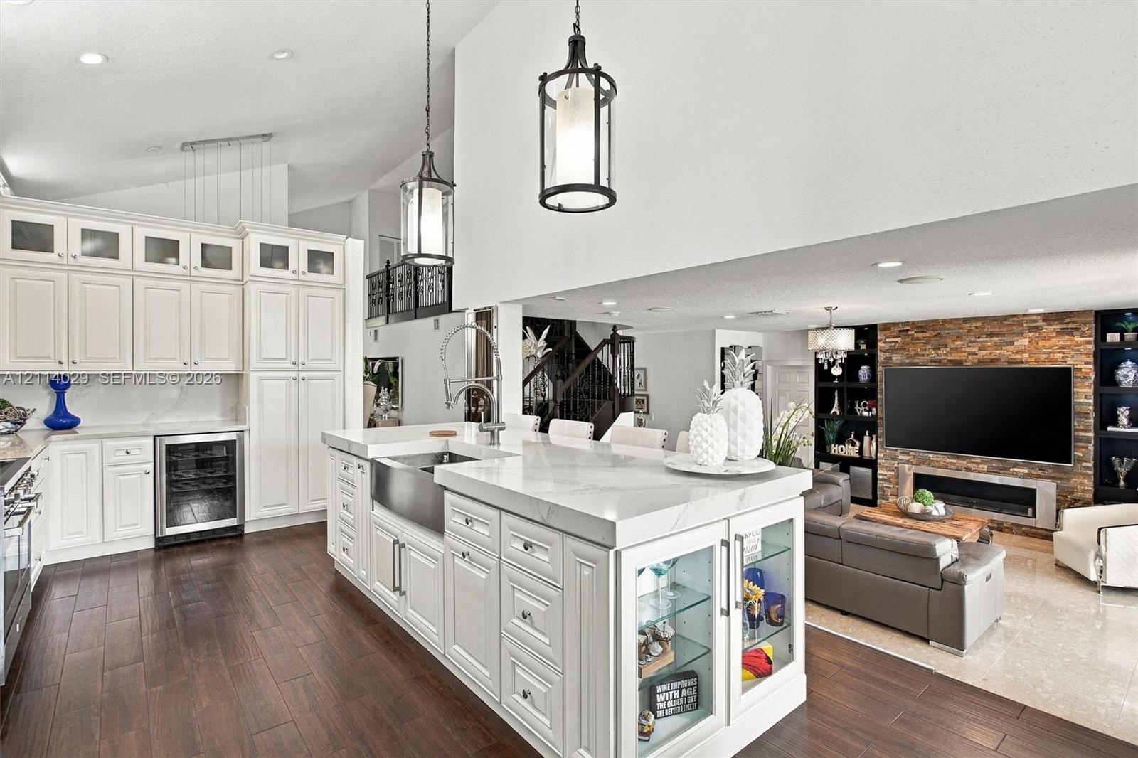 Kitchen featuring white shaker-style cabinetry, an expansive center island with an integrated farmhouse sink, glass-front display shelving, stone-veneer fireplace surround, and dark wood-finish flooring