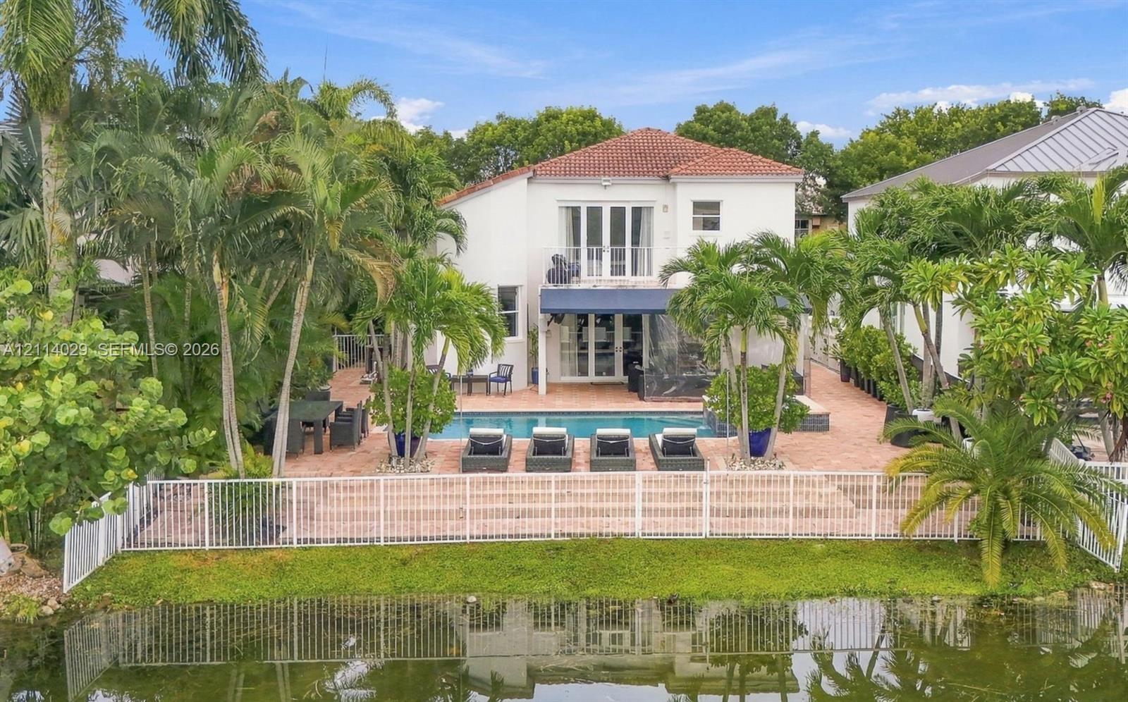 Waterfront rear exterior featuring a swimming pool, expansive brick-paver patio, second-story balcony, and mature tropical landscaping