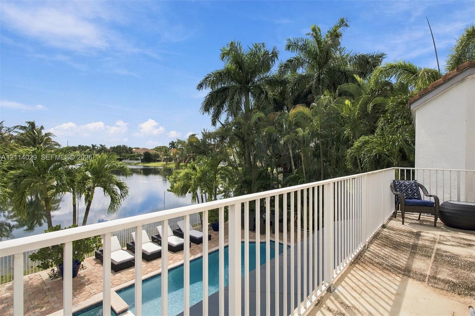 Second-story balcony with white metal railings overlooking a rectangular swimming pool, paver patio, mature palm trees, and a pond