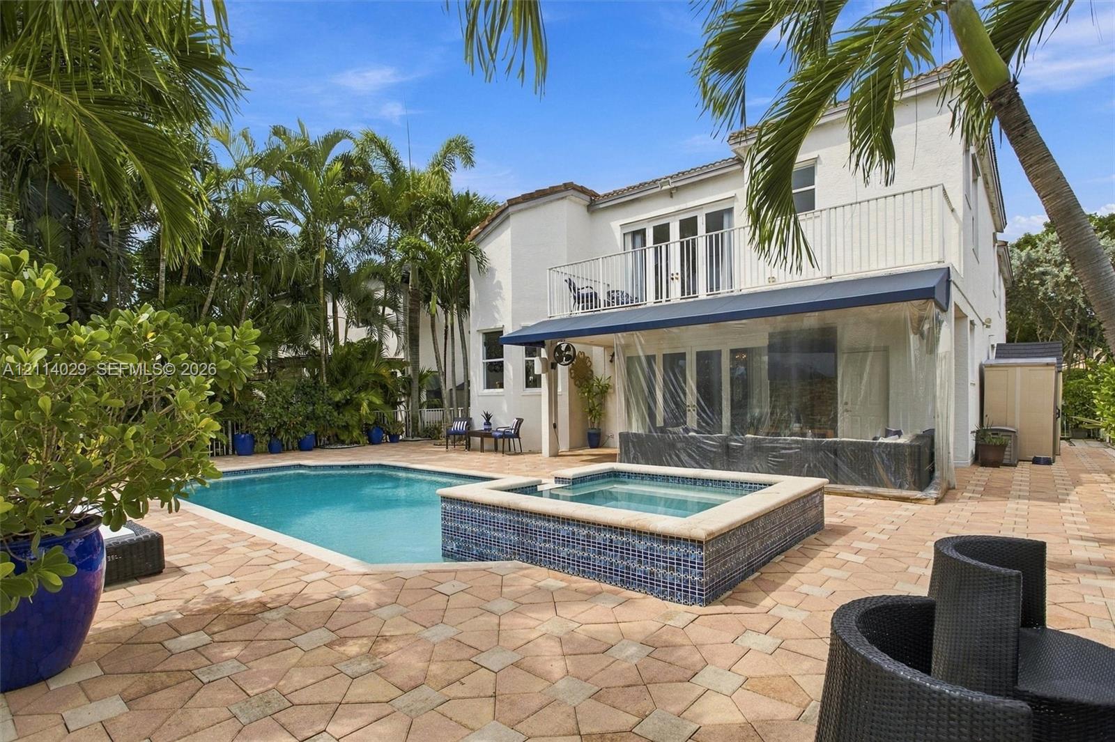 Exterior features including a swimming pool with a raised tiled spa, expansive paver patio, second-story balcony with metal railing, and retractable exterior awning