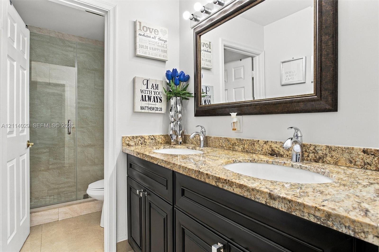 Dual-sink vanity with stone countertop, dark-toned cabinetry, framed wall mirror, multi-bulb vanity light, and tiled walk-in shower with glass enclosure