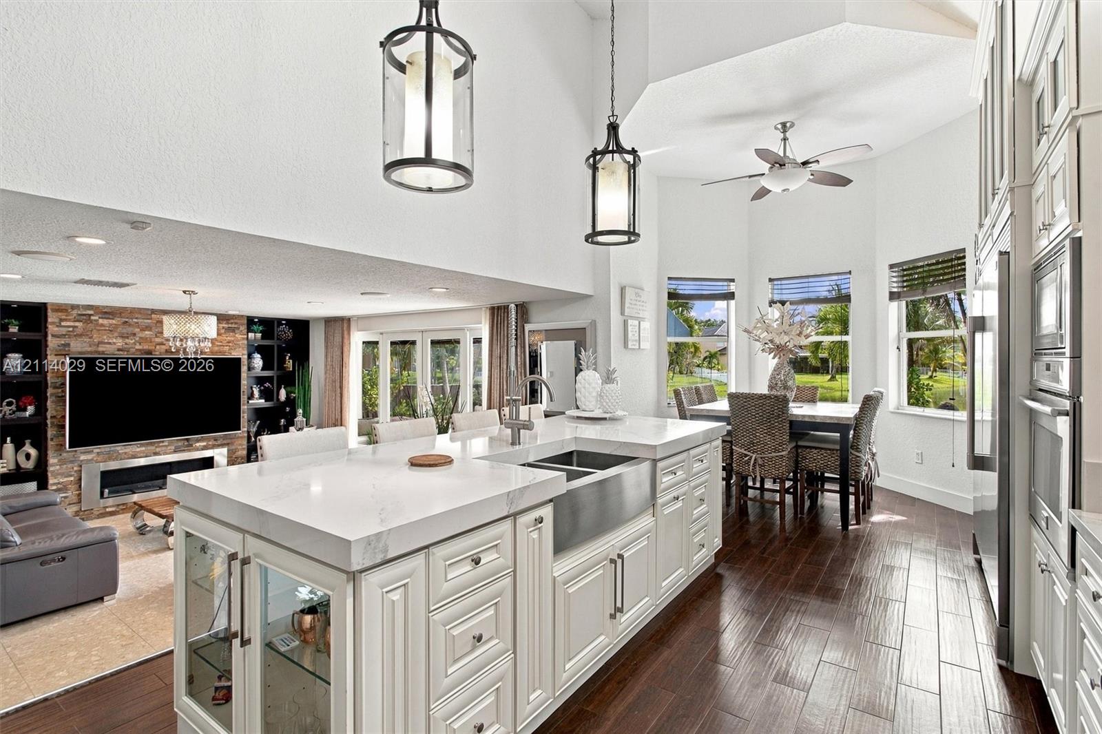 Kitchen featuring a central island with a quartz countertop, stainless steel apron-front sink, white cabinetry with glass-fronted accents, wood-finish flooring, and vaulted ceilings with hanging pendant lighting