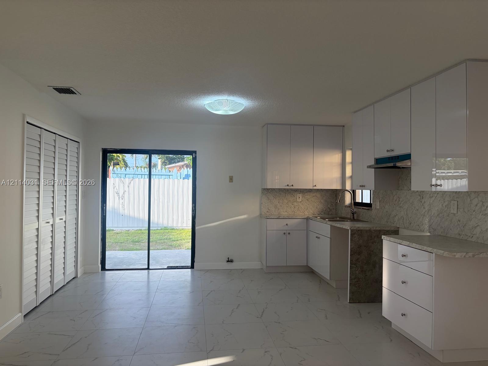 Kitchen featuring high-gloss white cabinetry, stone-patterned countertops, coordinating backsplash, large-format tile flooring, and sliding glass doors Kitchen featuring high-gloss white cabinetry, stone-patterned countertops, coordinating backsplash, large-format tile flooring, and sliding glass doors