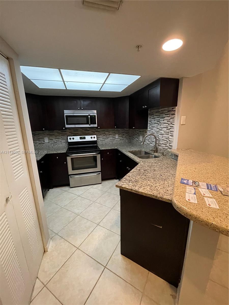 Kitchen featuring dark cabinetry, stone-finish countertops, stainless steel appliances, a multi-tone tile backsplash, and tile flooring