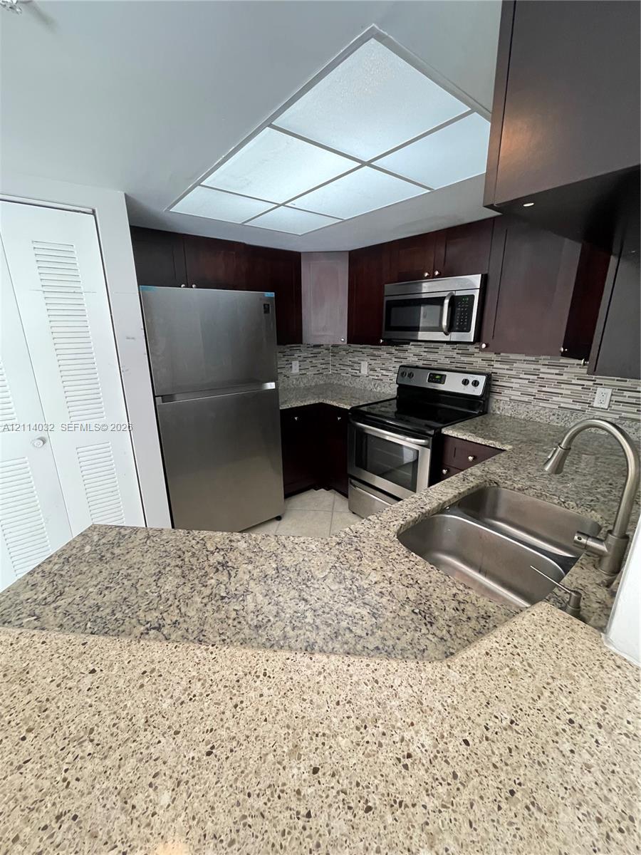 Kitchen featuring dark wood-finish cabinetry, speckled stone-composite countertops, stainless steel appliances, a multi-textured linear tile backsplash, and a double-basin stainless steel sink with a pull-down faucet