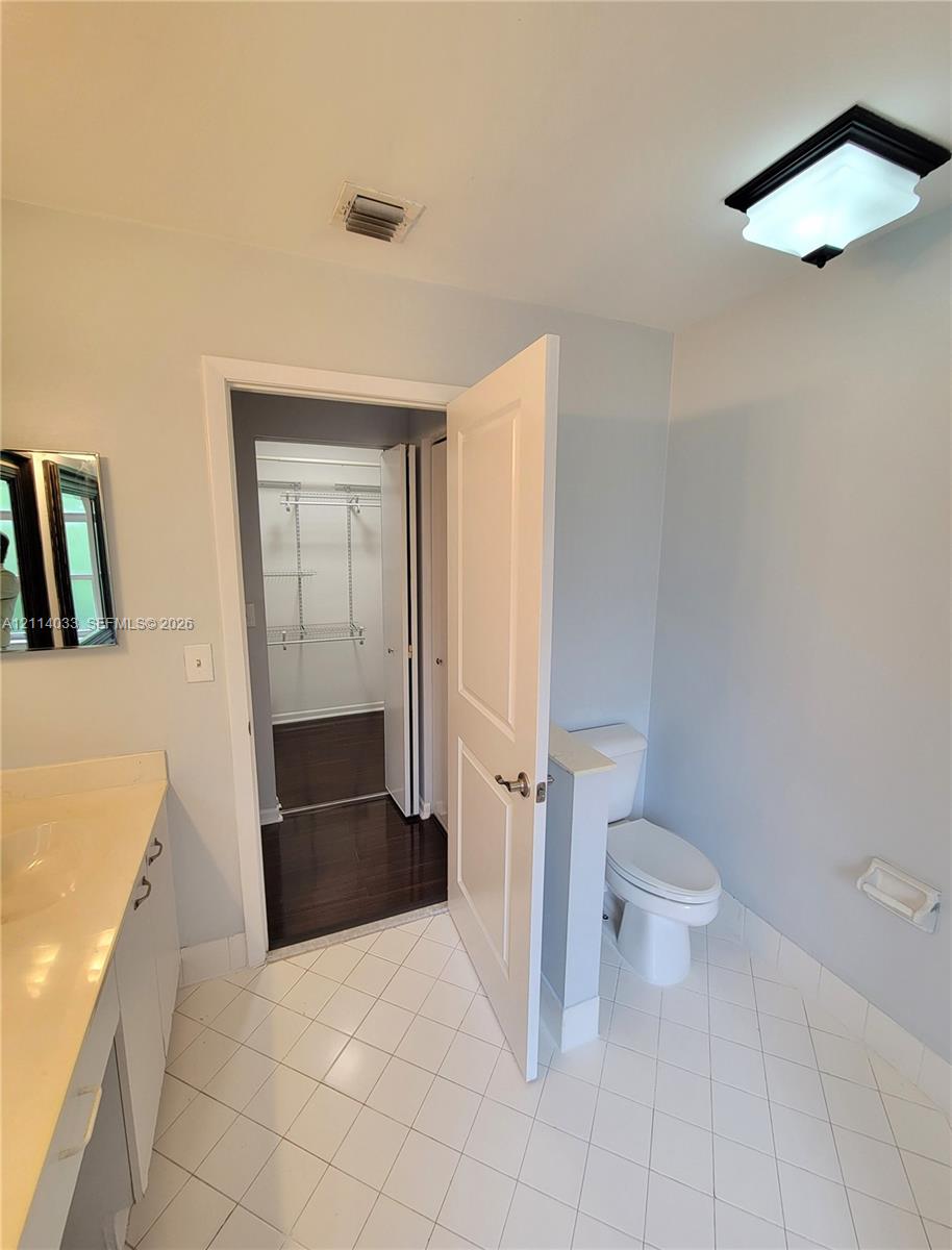 Bathroom featuring a vanity with integrated sink and countertop, white tile flooring, enclosed water closet with half-wall, and walk-in closet with wire shelving and dark wood-finish flooring