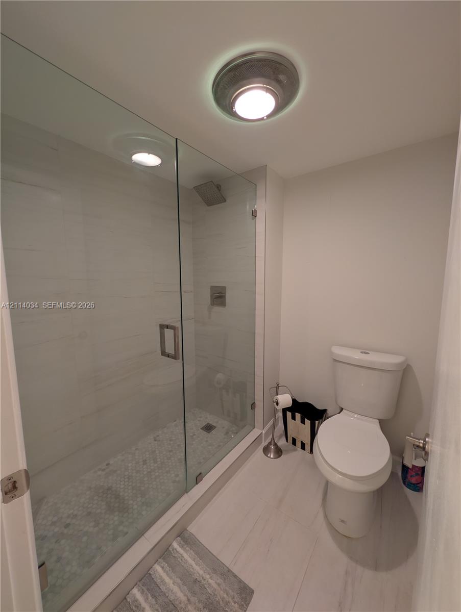 Walk-in shower featuring a glass enclosure, light-toned wall tile, and a decorative pebble-style floor, paired with a porcelain commode