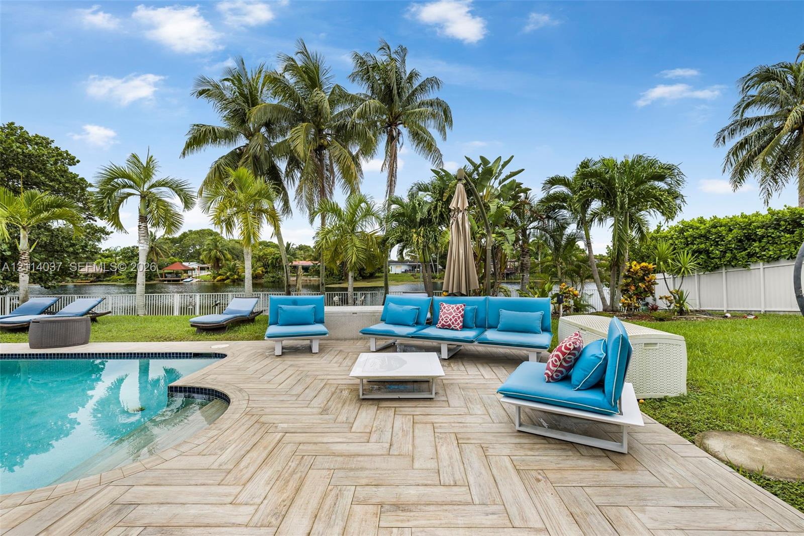 Waterfront backyard featuring a swimming pool with a stepped entry, herringbone-patterned tile decking, mature palm trees, and a white privacy fence