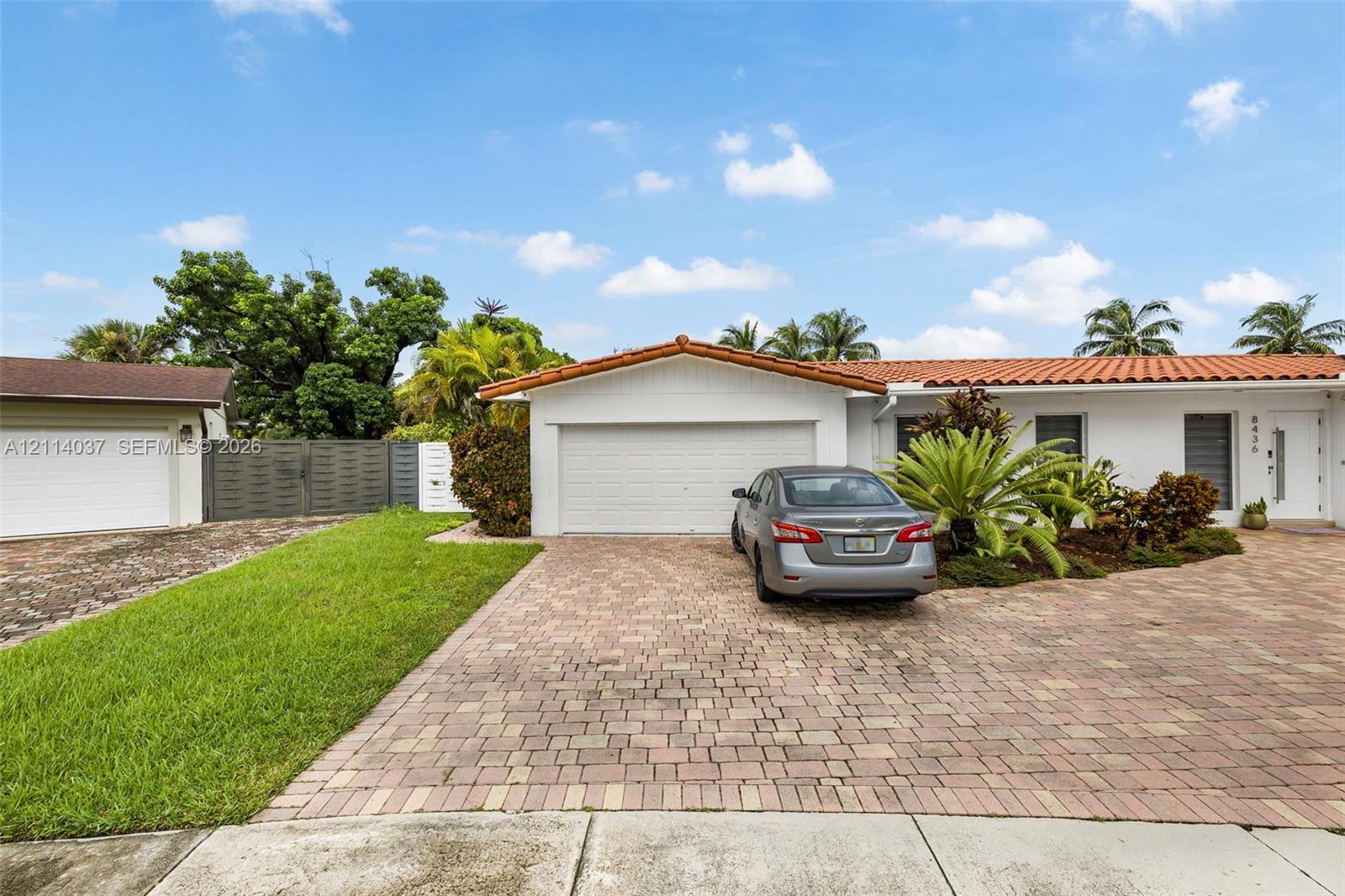 Single-story residence featuring a terracotta-tiled roof, white stucco exterior, paved brick driveway, two-car garage, and manicured landscaping