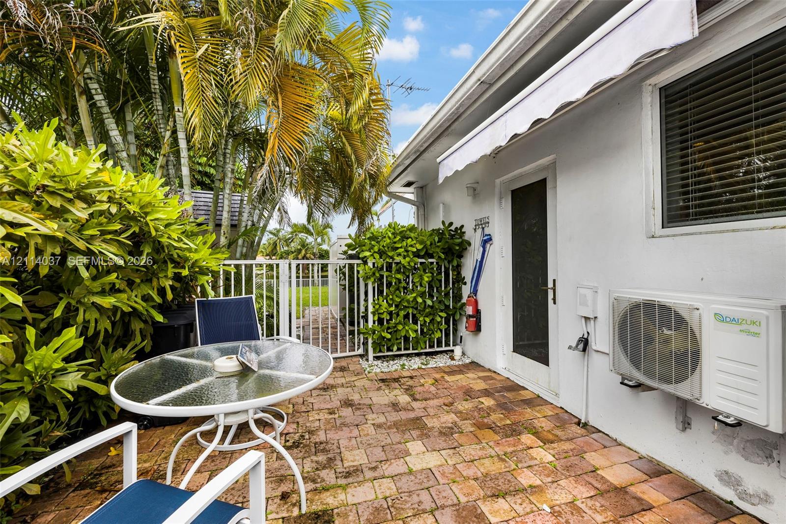Private patio featuring brick pavers, a retractable awning, and mature palm landscaping with a metal perimeter fence