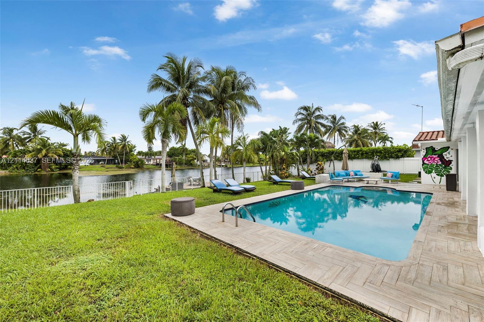Backyard featuring an in-ground swimming pool with herringbone-patterned stone decking, grass lawn, water frontage, and mature palm trees