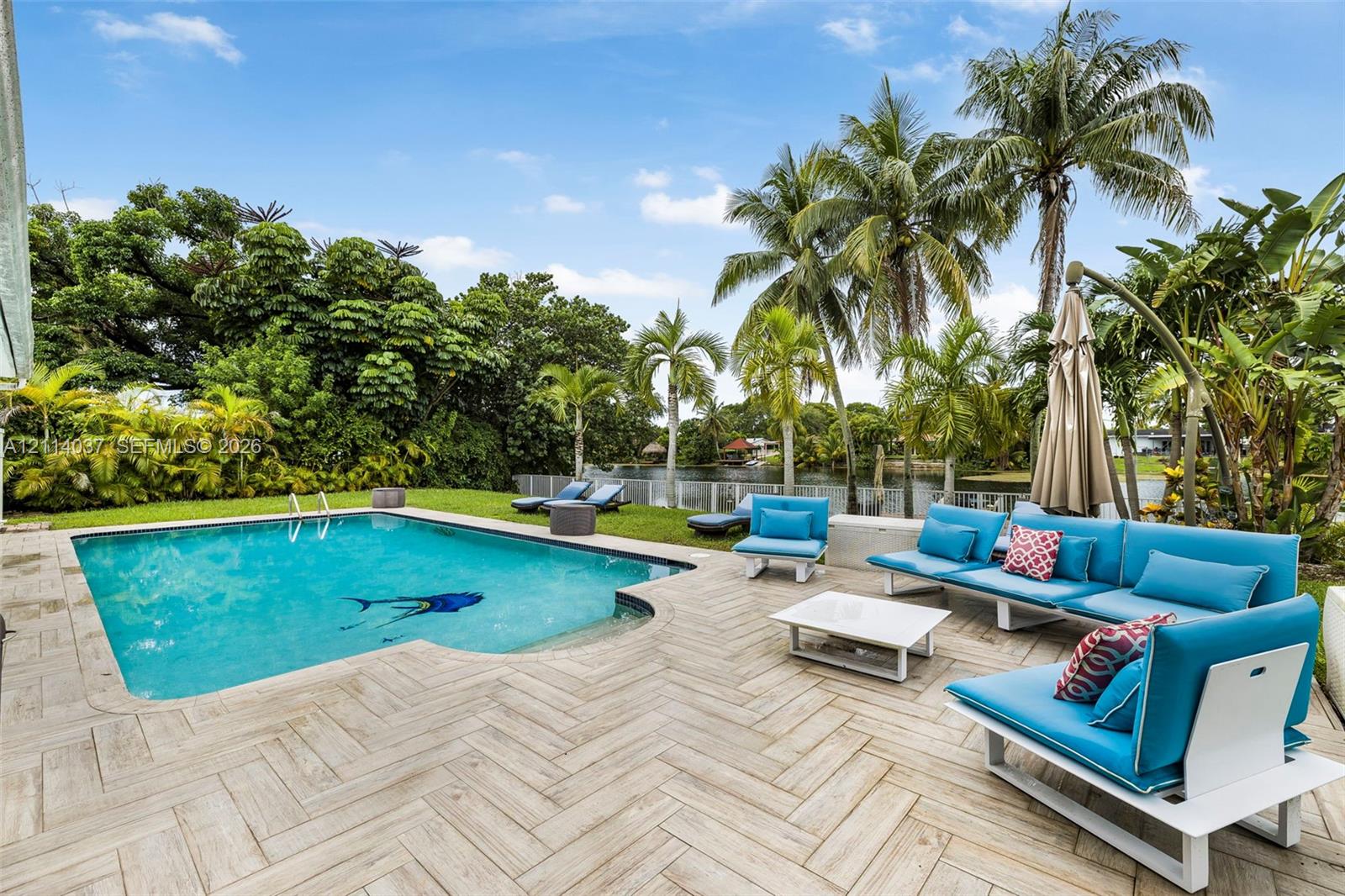 In-ground swimming pool with a herringbone-patterned tile deck, manicured lawn, and mature palm trees along the waterfront