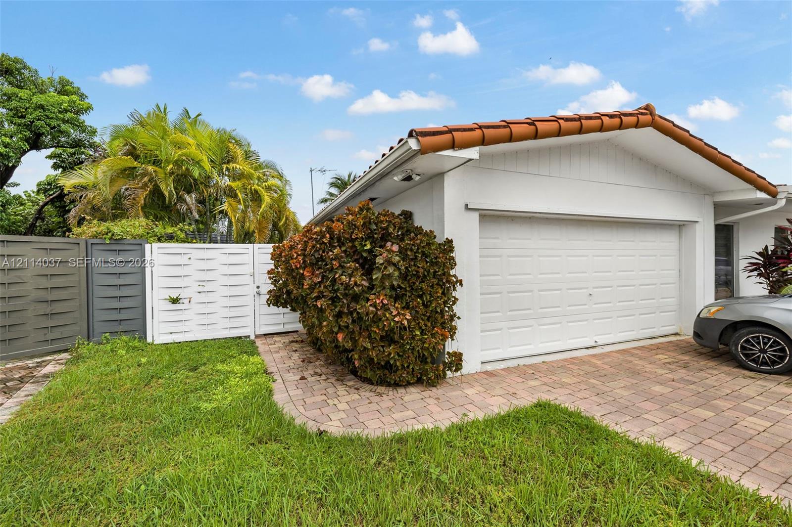 Exterior of a structure featuring a tiled roof, multi-panel garage door, and brick-paver driveway, with white privacy fencing and perimeter landscaping
