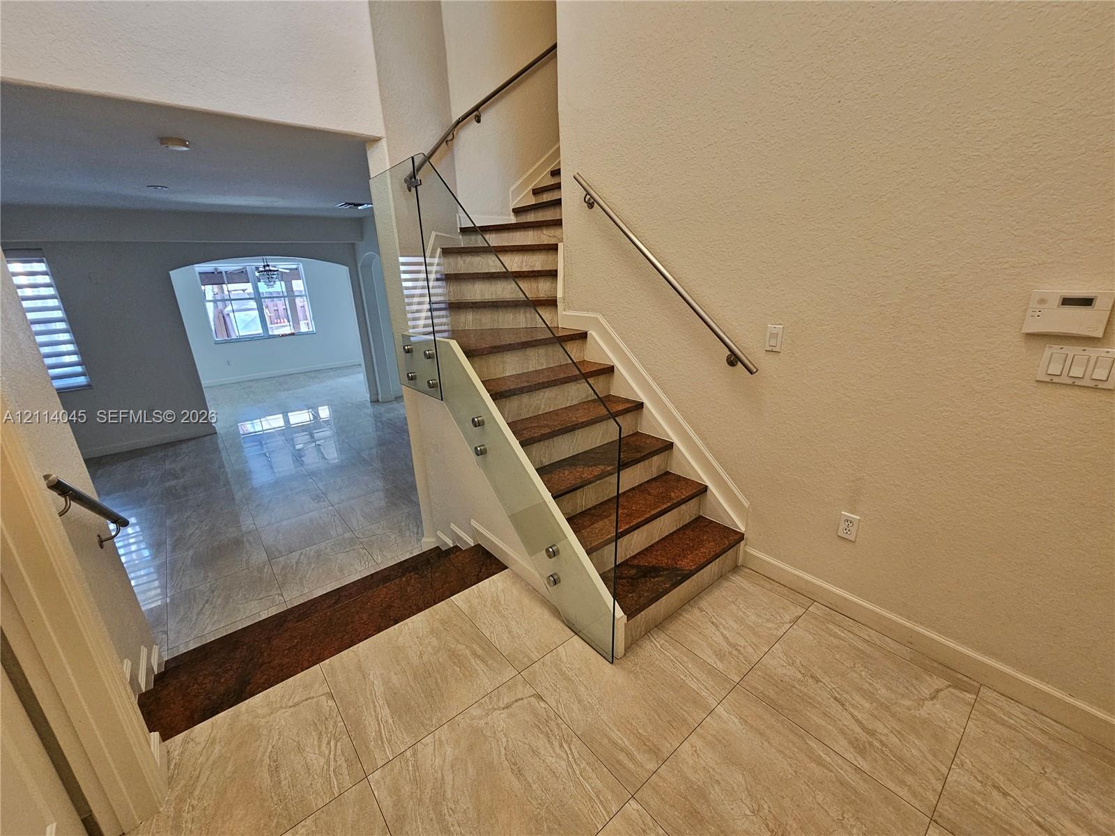 Staircase featuring wood-finish treads, glass panel railing with metallic standoffs, and a wall-mounted metal handrail, complemented by neutral tile flooring