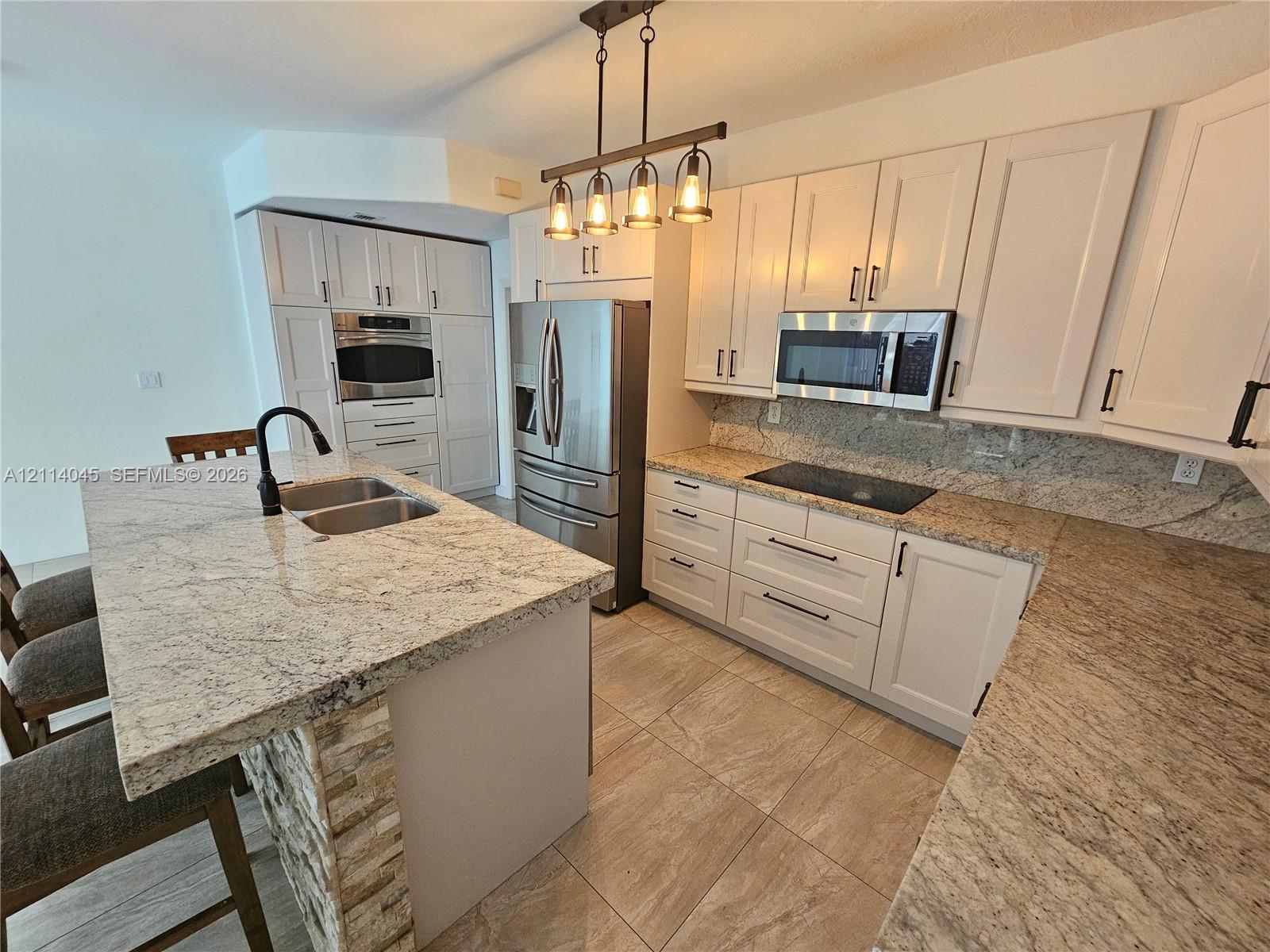 Kitchen featuring shaker-style cabinetry, stone countertops, stainless steel appliances, stone-accented center island with an undermount sink, and tile flooring