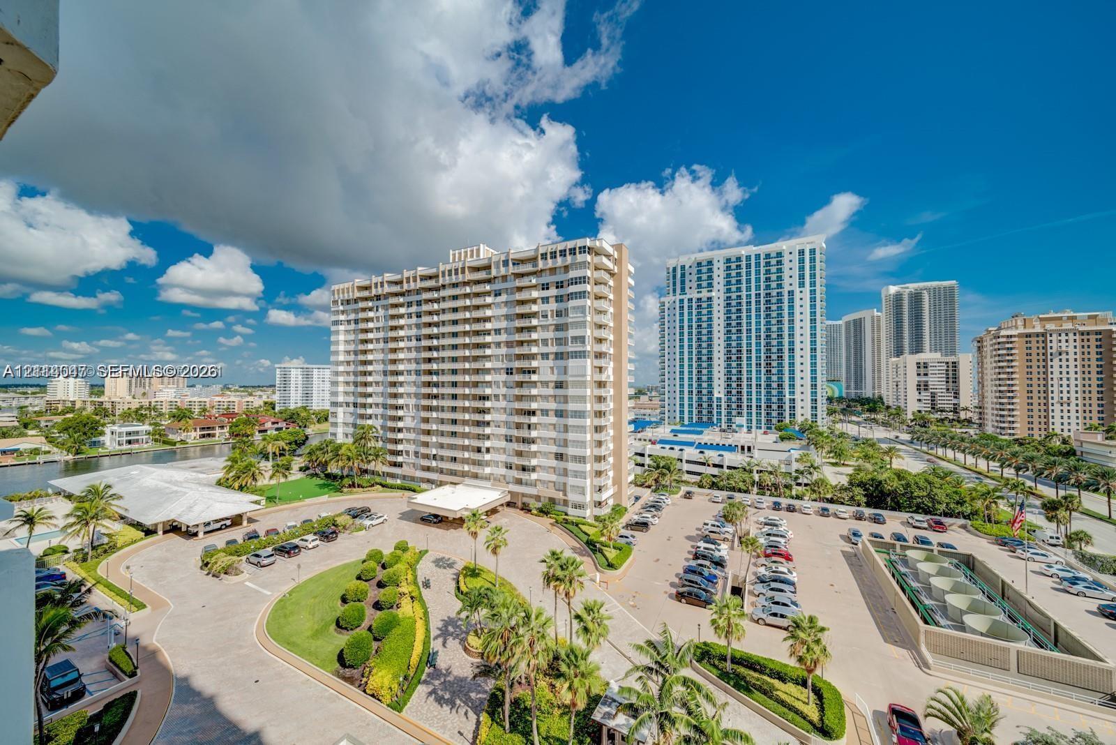 Elevated perspective featuring manicured landscaping, paved driveways, surface parking, and coastal building architecture