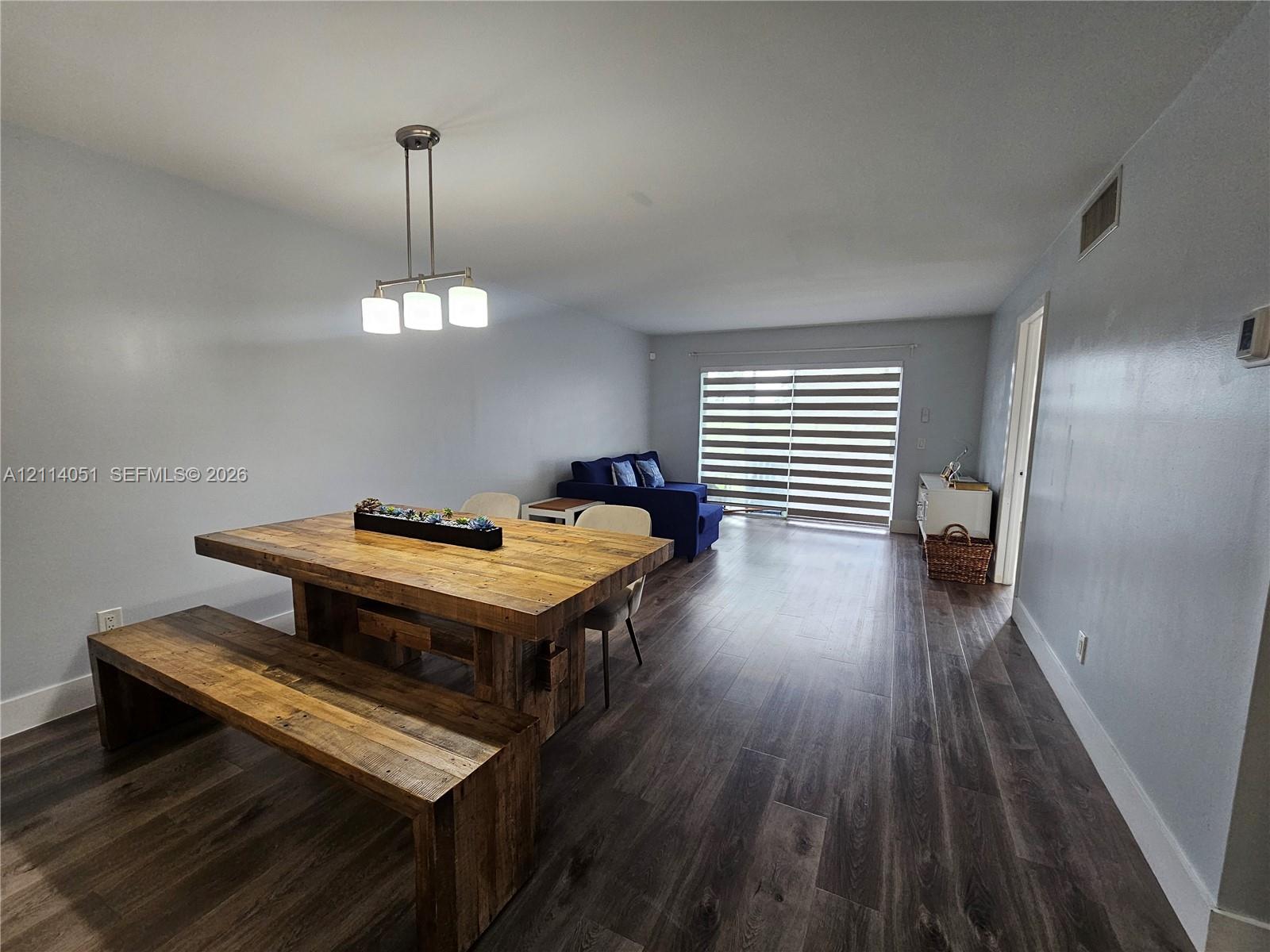 Open-concept interior featuring wood-finish flooring, a three-light pendant fixture, and sliding glass doors with horizontal blinds Open-concept interior featuring wood-finish flooring, a three-light pendant fixture, and sliding glass doors with horizontal blinds
