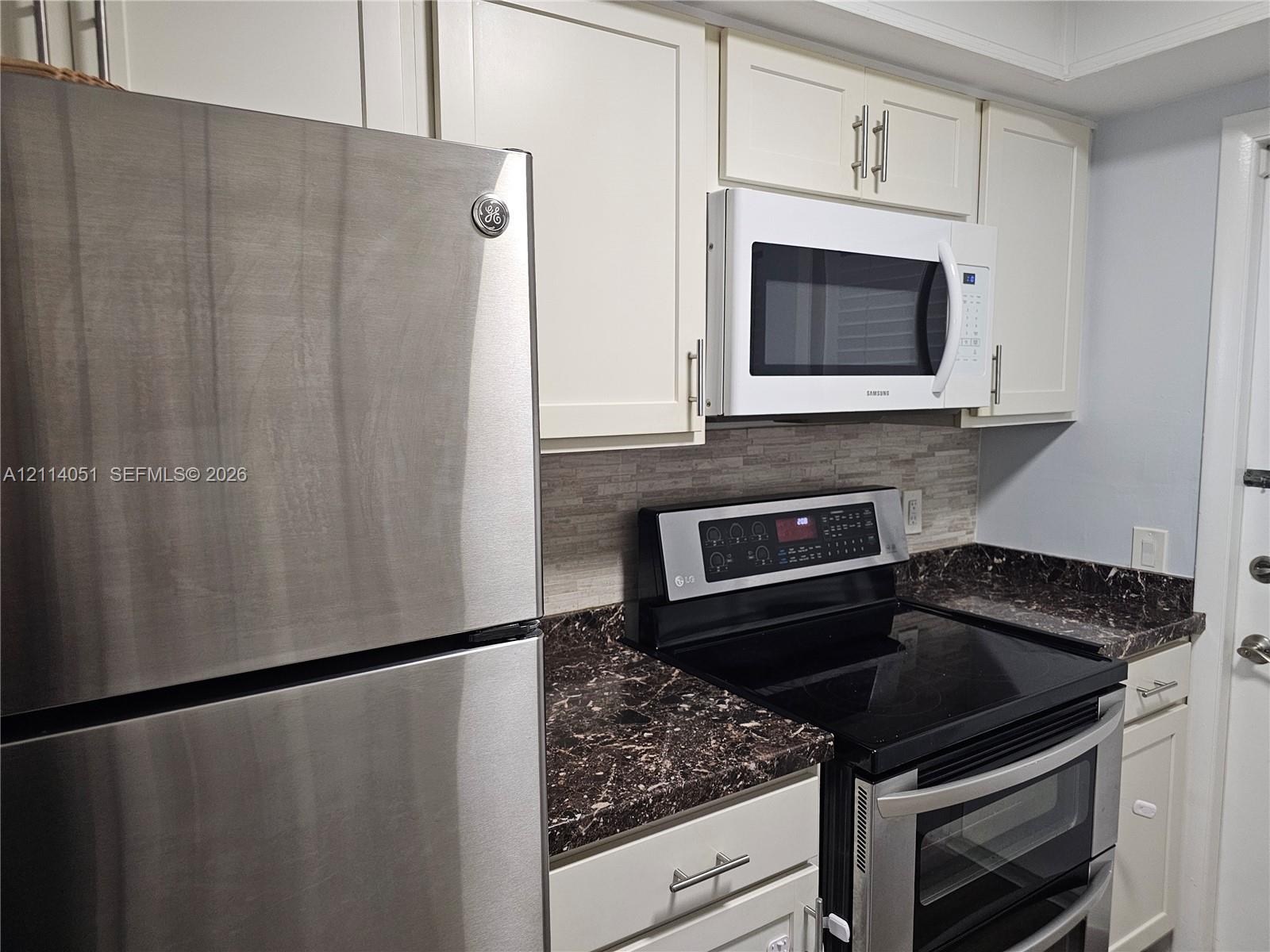 Kitchen featuring white shaker-style cabinetry, stainless steel refrigerator, dark stone-patterned countertops, stacked-tile backsplash, and integrated microwave Kitchen featuring white shaker-style cabinetry, stainless steel refrigerator, dark stone-patterned countertops, stacked-tile backsplash, and integrated microwave