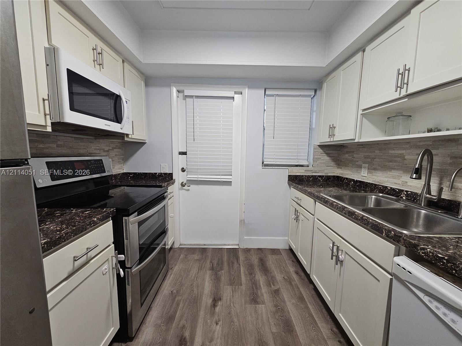 Kitchen featuring shaker-style cabinetry, dark stone-finish countertops, tiled backsplash, stainless steel double-basin sink, and wood-finish flooring Kitchen featuring shaker-style cabinetry, dark stone-finish countertops, tiled backsplash, stainless steel double-basin sink, and wood-finish flooring