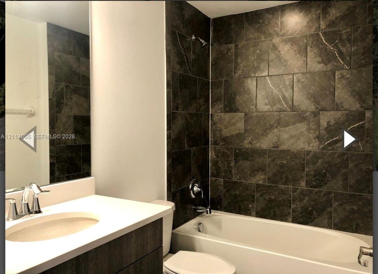 Bathroom featuring a vanity with white countertop, integrated sink, large-format dark stone-finish wall tiles surrounding the bathtub, and wall-mounted mirror Bathroom featuring a vanity with white countertop, integrated sink, large-format dark stone-finish wall tiles surrounding the bathtub, and wall-mounted mirror