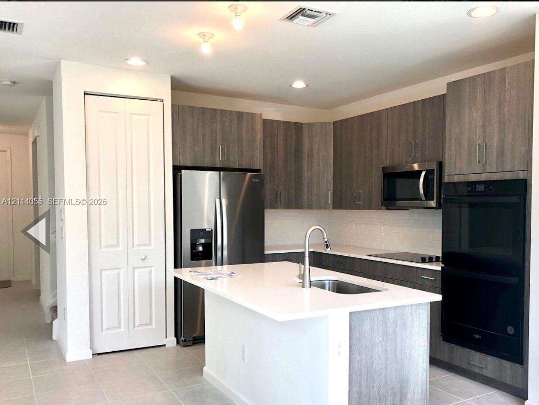 Kitchen featuring wood-finish cabinetry, quartz countertops, central island with stainless steel sink, suite of built-in stainless steel appliances, and tile flooring Kitchen featuring wood-finish cabinetry, quartz countertops, central island with stainless steel sink, suite of built-in stainless steel appliances, and tile flooring